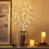 A vase with glowing LED branches beside lit candles and a framed painting on a shelf. | 28Alchemy