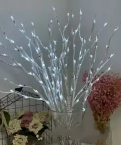 White LED twig lights in a glass vase, with pink dried flowers and a bouquet of roses on a table in the background. | 28Alchemy