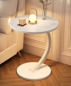 Round white side table with curved base, holding a candle, small lamp, and vase with flowers beside a beige sofa. | 28Alchemy