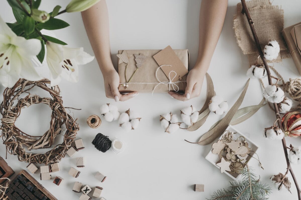 Hands wrapping a gift with brown paper surrounded by natural decorations, twine, and craft supplies on a white table. | 28Alchemy