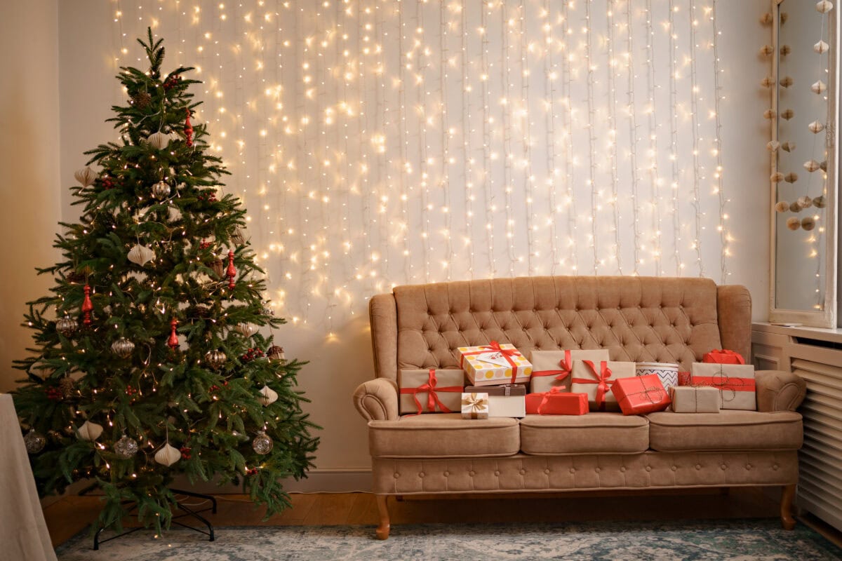A Christmas tree and a brown sofa with wrapped gifts in a cozy, well-lit living room decorated with string lights. | 28Alchemy