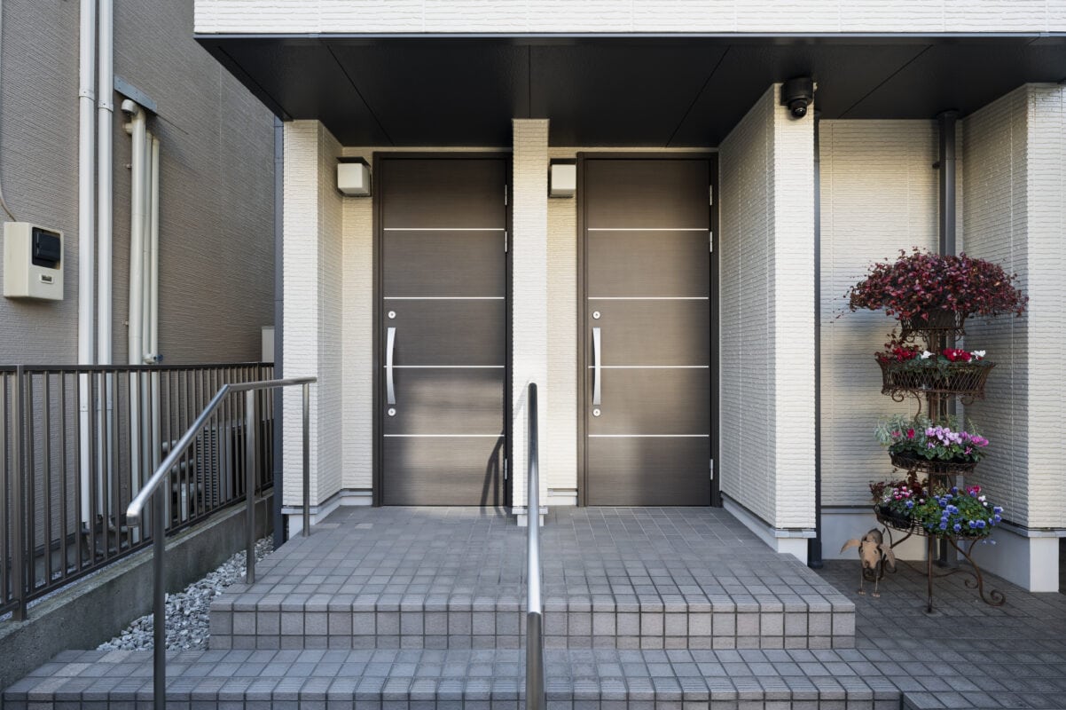 Two modern front doors with steps, metal railing, and a tiered flower stand with colorful plants on the right side. | 28Alchemy
