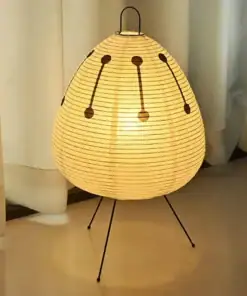 A glowing paper lantern lamp with black tripod legs stands on a floor in front of white curtains. | 28Alchemy