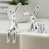 Two shiny silver rabbit figurines with long ears, posed playfully on a white surface with a plant in the background. | 28Alchemy