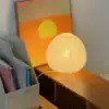 A round paper lamp glows on a wooden table next to books and a painting of an orange sunset with a yellow sun. | 28Alchemy