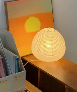 A round paper lamp glows on a wooden table next to books and a painting of an orange sunset with a yellow sun. | 28Alchemy