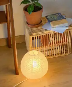 A round paper lamp glows on the floor beside a wicker basket, a potted plant, and stacked books. | 28Alchemy