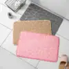 Three textured bath mats in pink, beige, and gray are laid on a white tiled floor near slippers and toiletries. | 28Alchemy