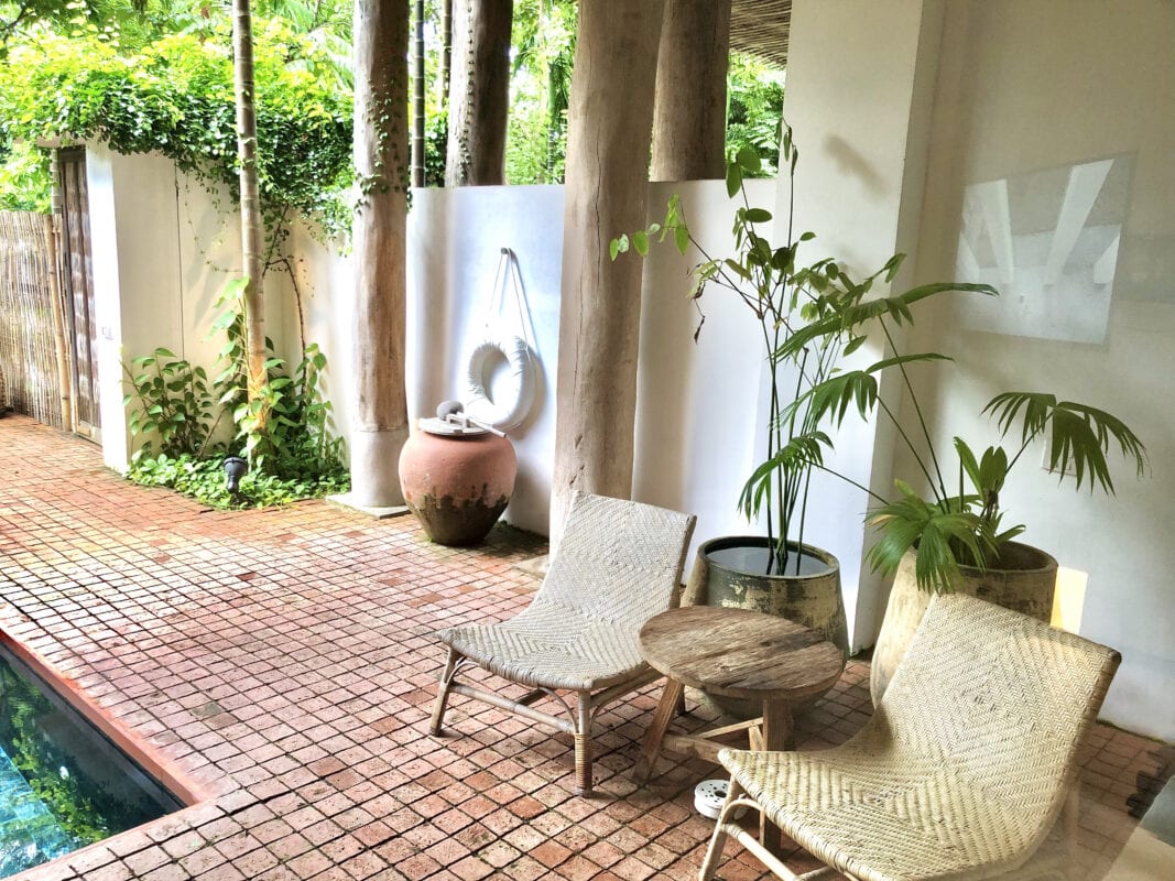 Two wicker chairs and a small table sit beside a tropical pool, surrounded by lush plants and tall trees. | 28Alchemy