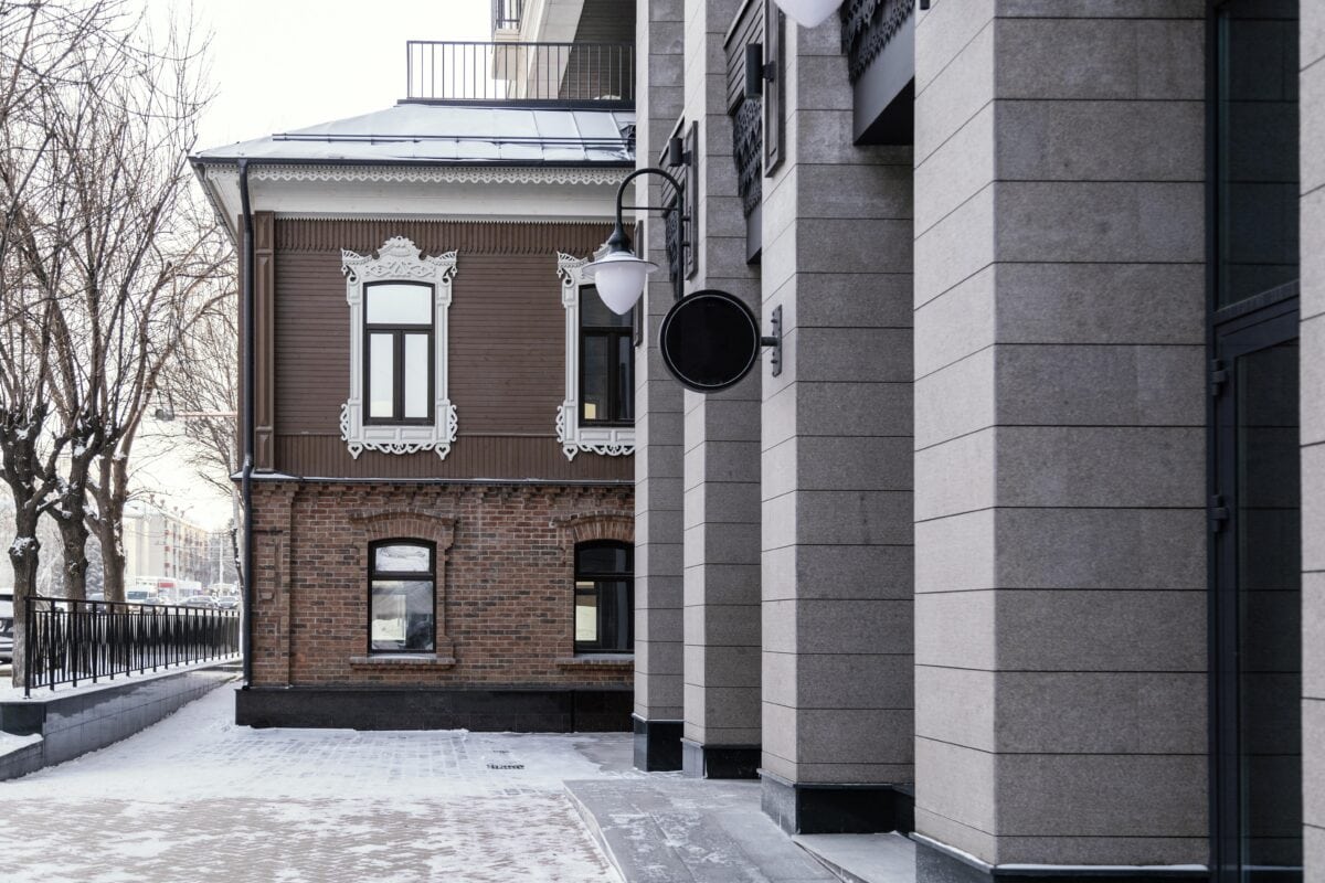 Modern stone building next to an old brick and wood building on a snowy street, with bare trees and street lamps. | 28Alchemy