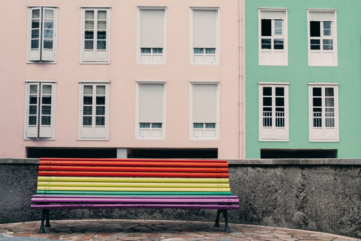 A rainbow-colored bench sits in front of pastel pink and green buildings with white shuttered windows. | 28Alchemy
