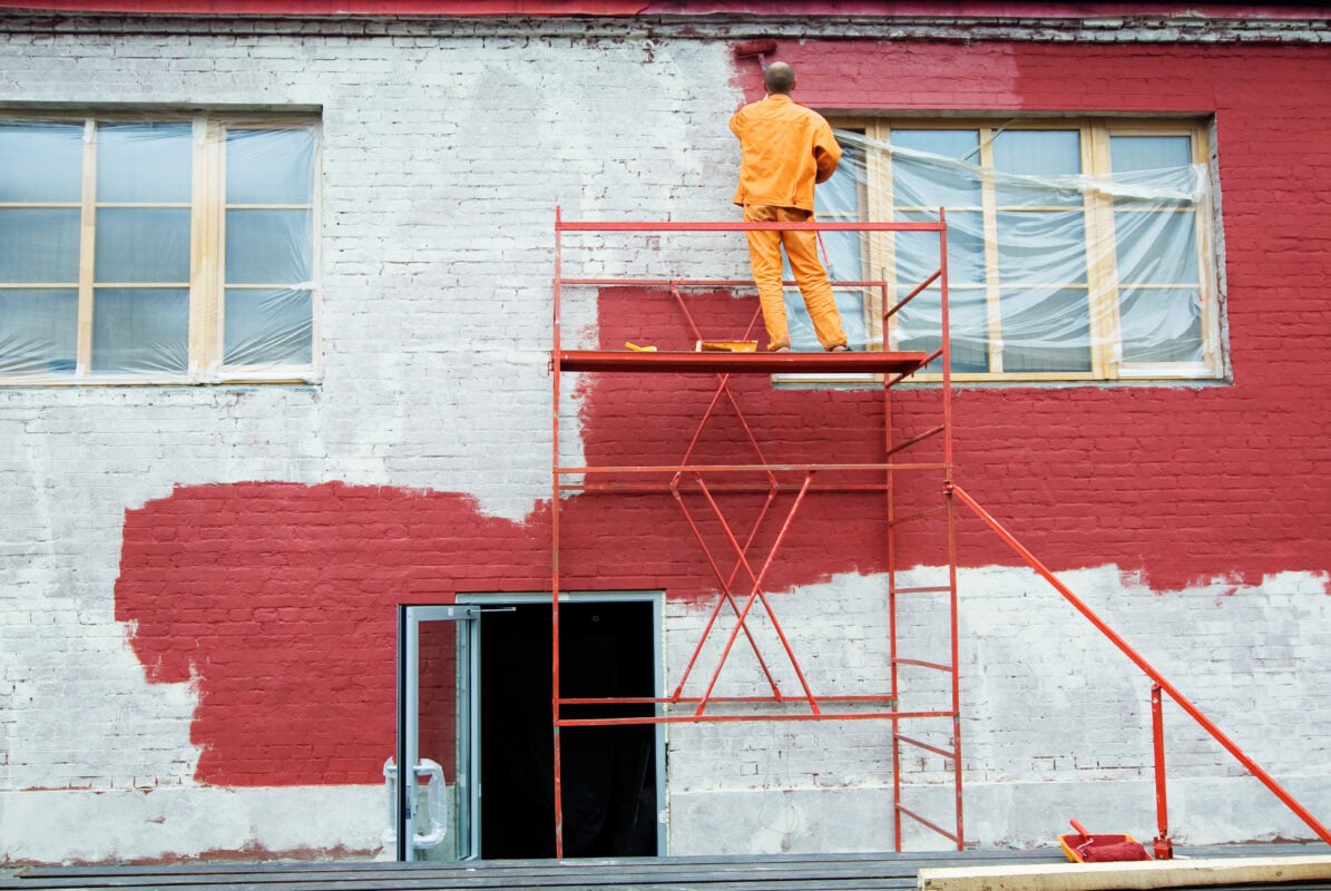 A person on scaffolding paints a white brick building red; windows are covered with plastic sheets. | 28Alchemy