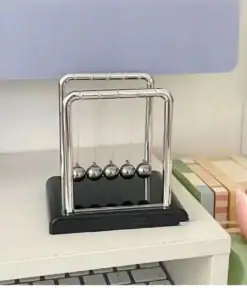 A Newton's cradle with five metal balls sits on a white desk beside a stack of colorful books. | 28Alchemy