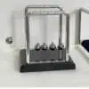 A Newton's cradle with one ball lifted, showing the classic metal balls on a black base. | 28Alchemy