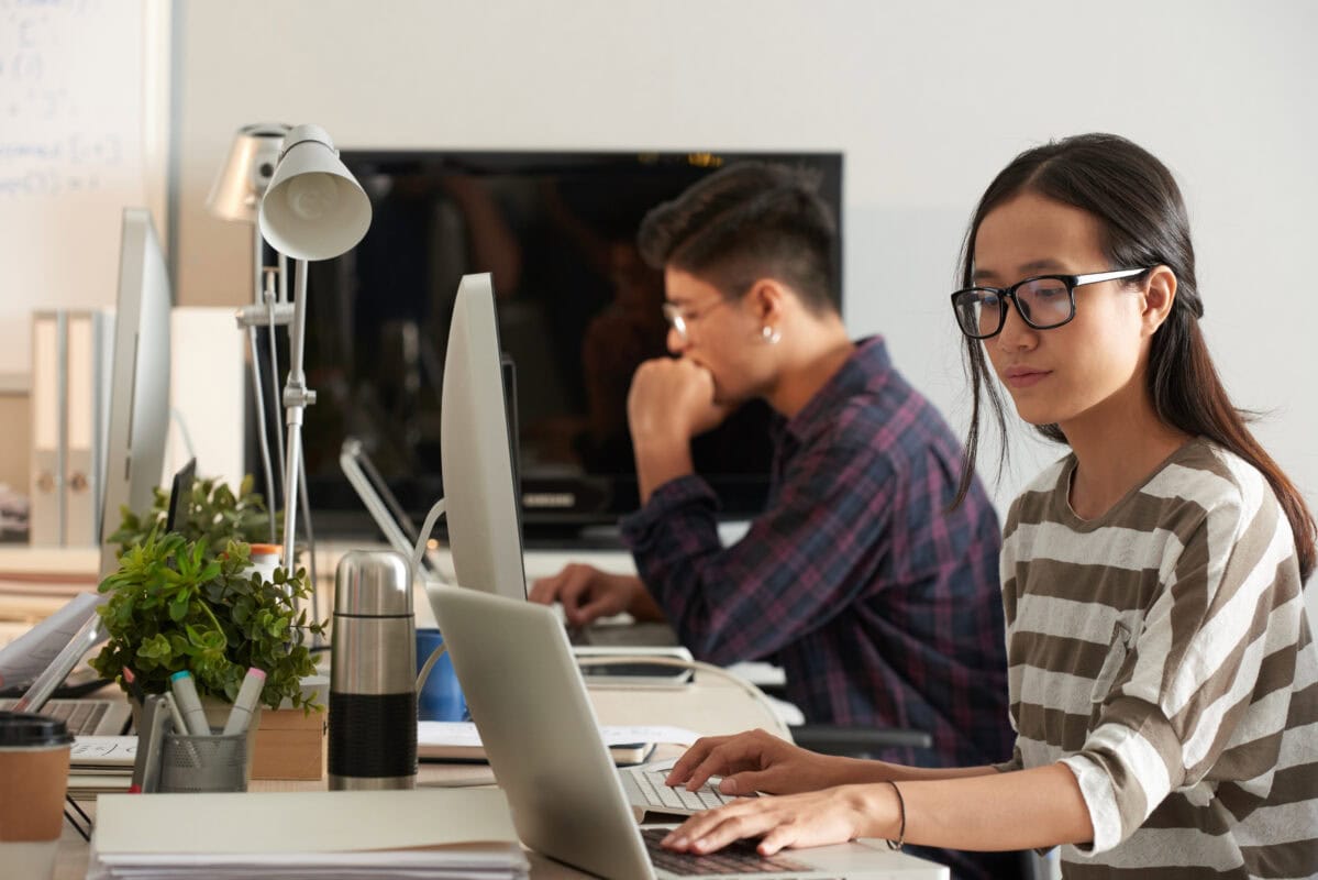 Two people working on computers at desks in a modern office, with plants and office supplies nearby. | 28Alchemy