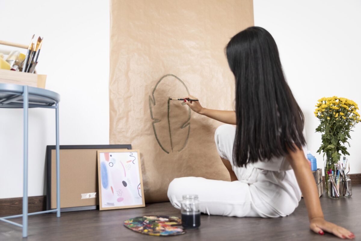 Woman with long dark hair sits on the floor sketching a face outline on brown paper; art supplies nearby. | 28Alchemy