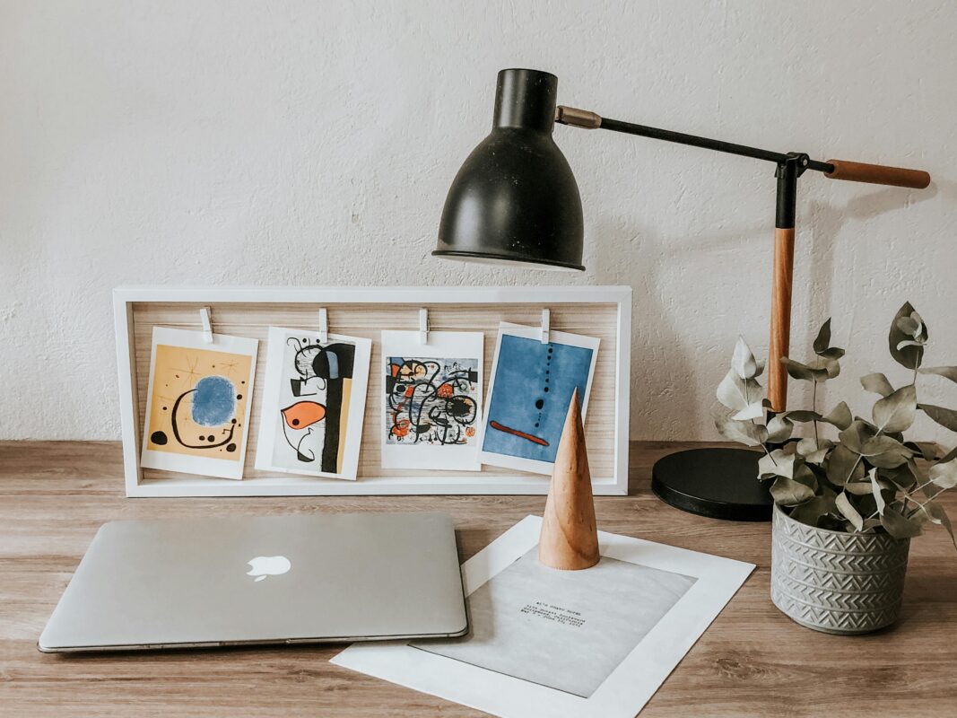 A desk with a laptop, lamp, plant, framed art postcards, and a wooden cone on a wooden surface. | 28Alchemy