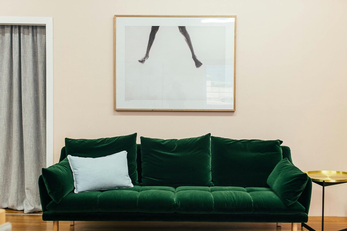 A green velvet sofa with a light blue pillow sits beneath a framed photo of legs on a pale wall, near a gold side table. | 28Alchemy