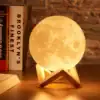 A moon-shaped lamp on a wooden stand, glowing warmly on a wooden table, with books and a brick wall in the background. | 28Alchemy