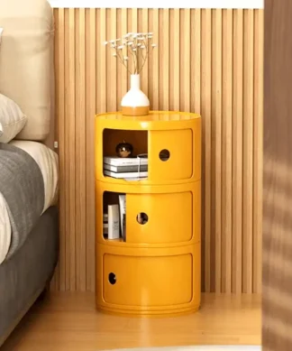 Yellow cylindrical nightstand with three shelves, displaying books, a clock, and a small white vase with flowers. | 28Alchemy