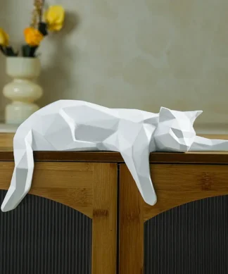 A geometric white cat sculpture lies stretched out on top of a wooden cabinet, with flowers in a vase in the background. | 28Alchemy