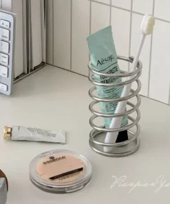 A tube of toothpaste and a toothbrush are in a spiral holder on a desk near a keyboard and makeup items. | 28Alchemy
