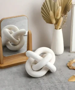 White knot sculpture reflected in a small wooden-framed mirror on a gray surface, with a white vase holding dried plants. | 28Alchemy