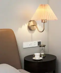Wall lamp above a bedside table with a teacup, saucer, hourglass, and power outlets on a light-colored wall. | 28Alchemy