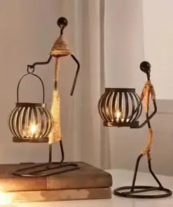 Two metal candle holders shaped like figures holding lanterns, with lit candles inside, placed on a table near books. | 28Alchemy