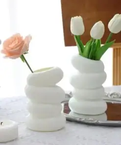 Two white, stacked, wavy vases hold a pink rose and three white tulips on a lace table with a mirror and candle. | 28Alchemy